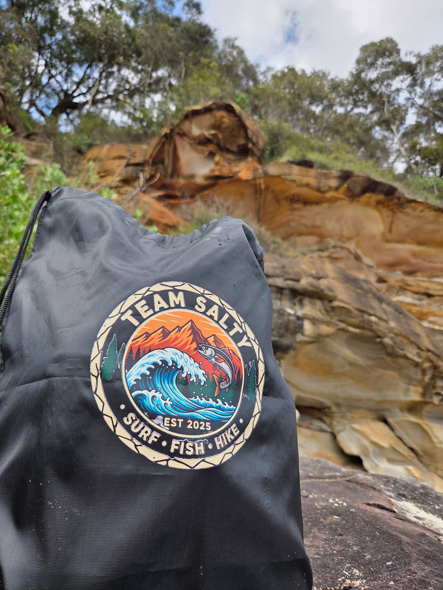 Black bag with a logo featuring a fish and text, set against a natural rock and tree background.